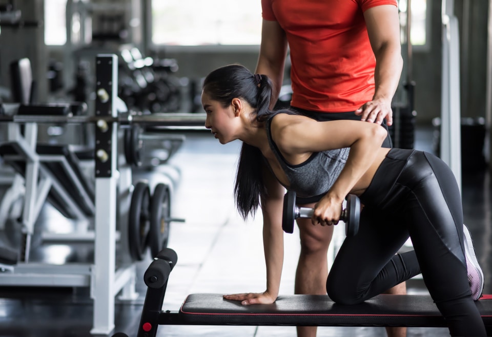 Jeune femme asiatique faisant un rowing avec haltère sur banc, coach masculin en rouge la supervise dans une salle de sport.